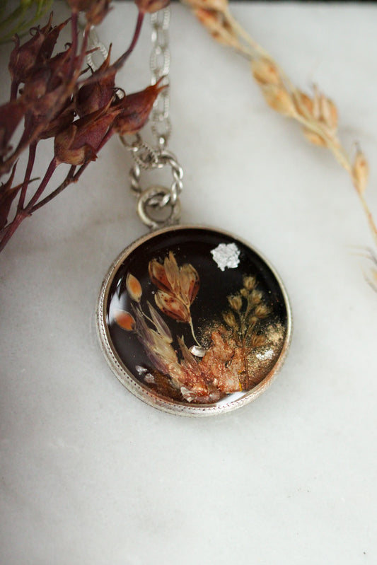 A round pendant necklace featuring a silver-toned bezel and chain, containing a black background with dried botanical elements, topped with resin