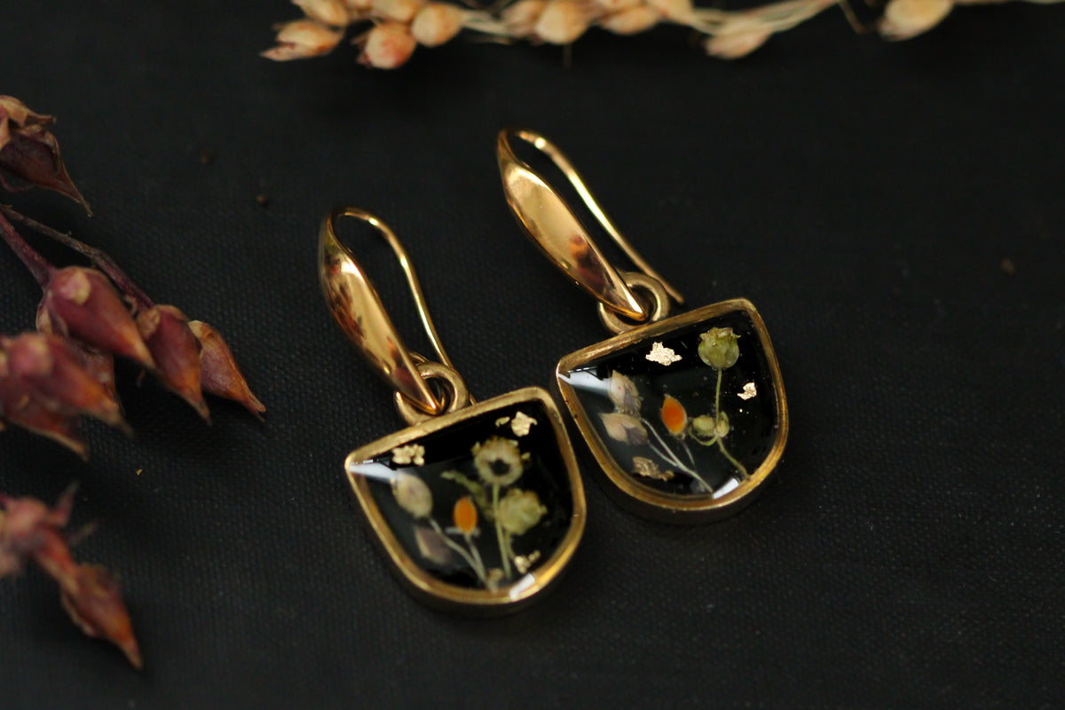 a pair of gold and black earrings featuring black background with dried plants and flowers topped with resin