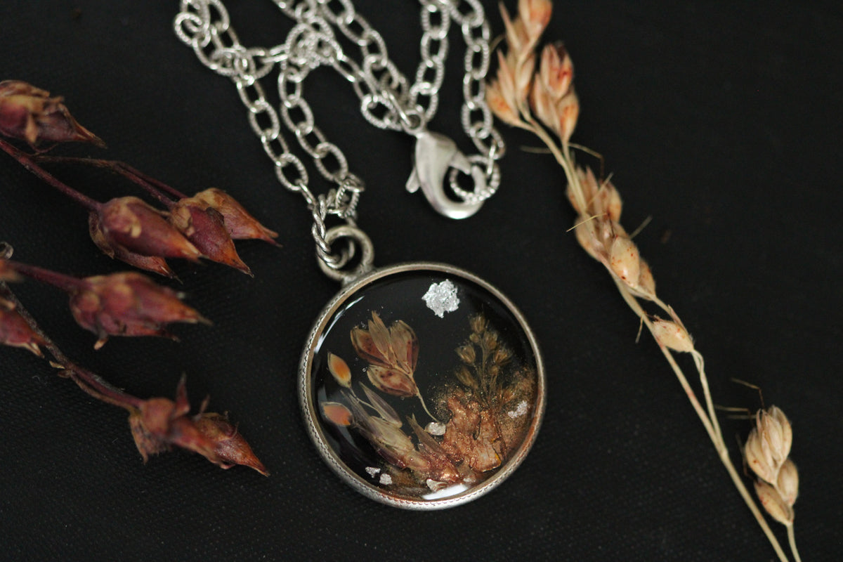 A round pendant necklace featuring a silver-toned bezel and chain, containing a black background with dried botanical elements, topped with resin