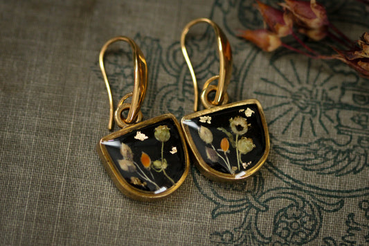 a pair of gold and black earrings featuring black background with dried plants and flowers topped with resin
